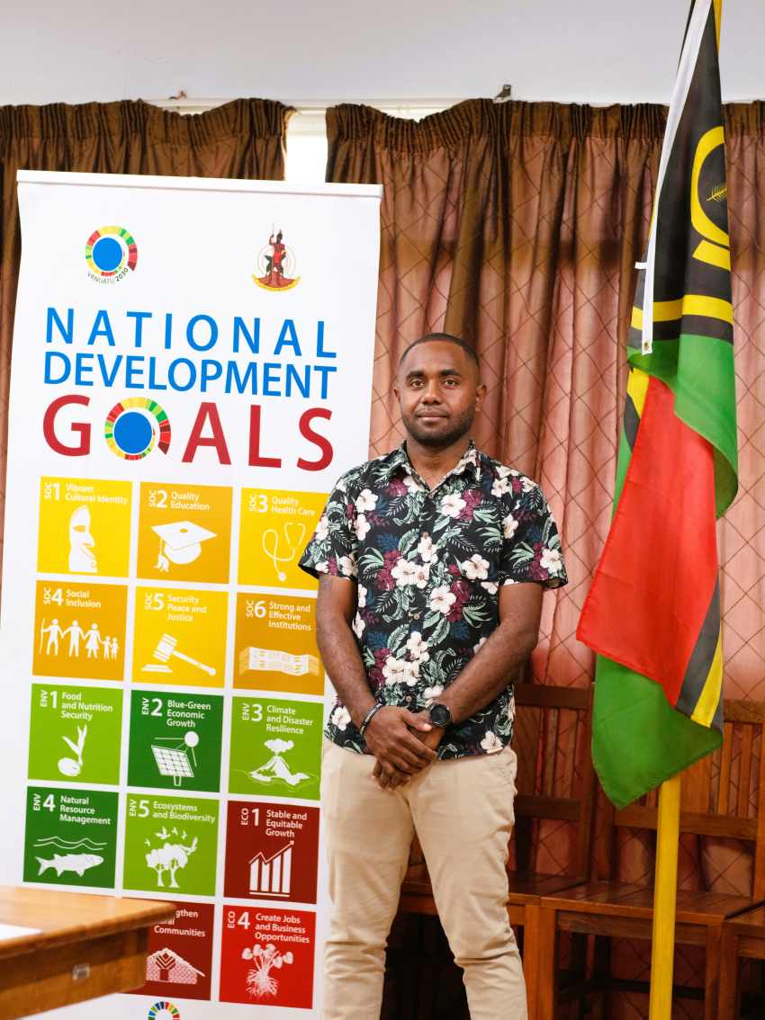 person stands in front of vanuatu flag and banner with national development goals