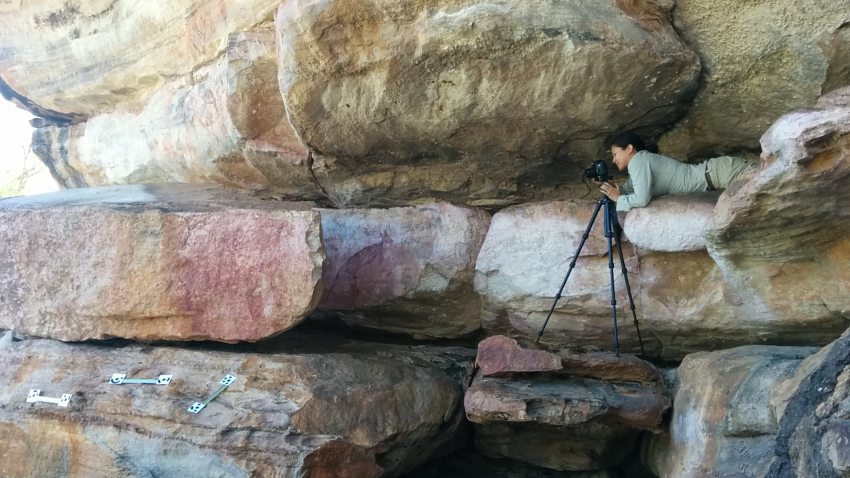 Photograph of a woman laying on a rock formation taking photographs of rock art