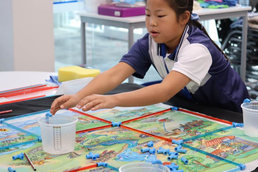 A young girl leans over a table covered in a map of a town, with blue and red plastic pipes laid out across it in a grid