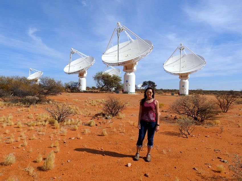 A woman stands on red dirt in the outback, with five white telescope dishes visible behind her in the distance