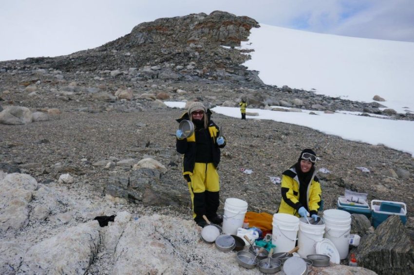 Photograph of two researchers in snow gear surrounded by equipment. They are pictured in a rocky area with snow in the distance