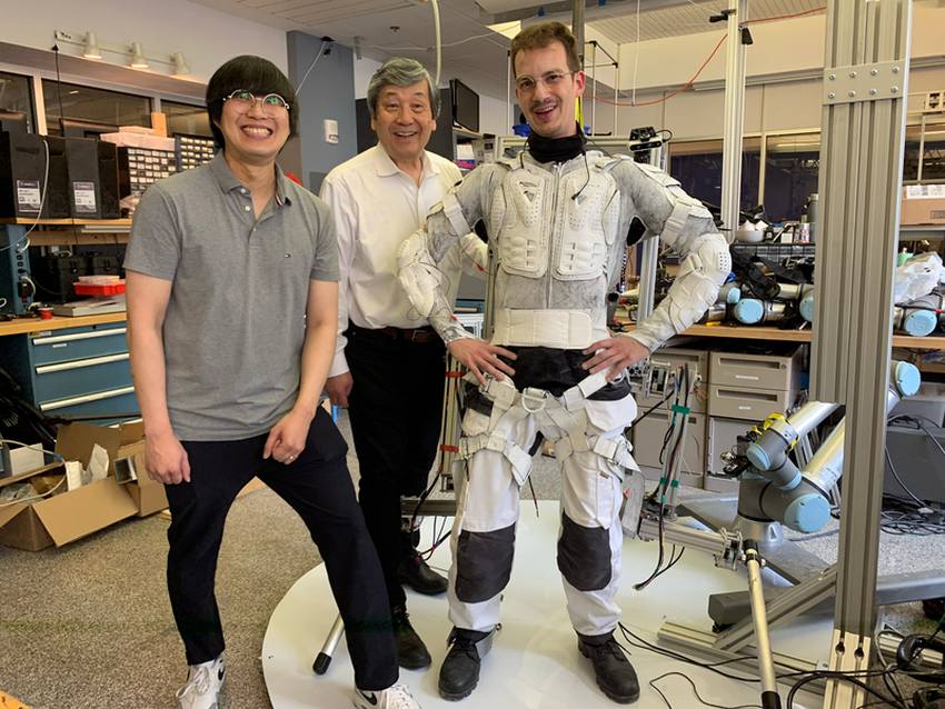 A photograph of 3 men inside a lab. Left is a young man in a grey polo shirt and black pants. Centre is an older man wearing  a white shirt and black dress pants. Right is a young man wearing a suit of white panels and straps. 2 metallic robot arms are protruding from something strapped behind his back.