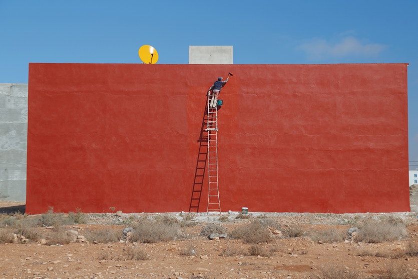 A man painting a wall red against a blue sky.