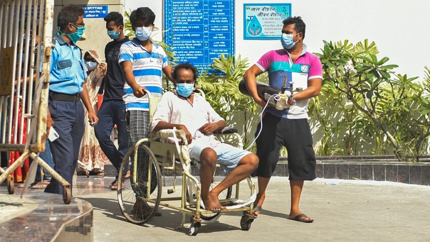 A covid-19 patient in a wheel chair with an oxygen cylinder. One man is pushing the chair and another is holding the oxygen tanks. A government official walks behind them