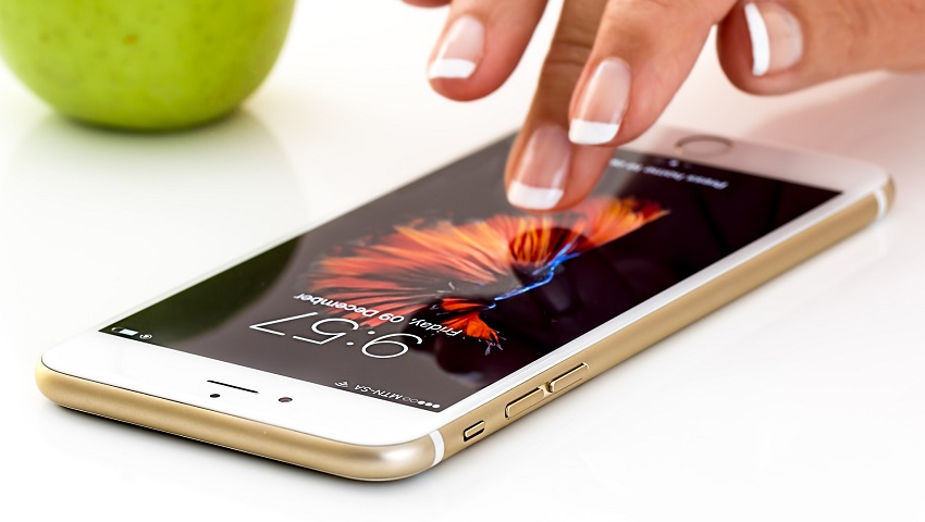 Fingers touching a smartphone screen. The time on the phone says 9:57. there is an apple in the background.