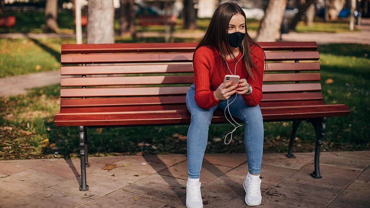 A woman sits on a park bench, wearing a mask and headphones.