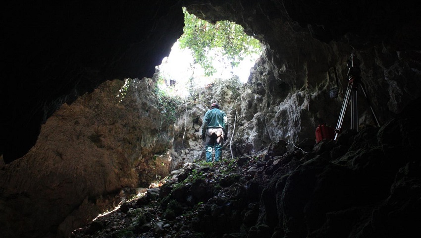 A cave with a hole in the middle. There are trees outside. a person stands next to the hole. Scientific equipment sits at the side.