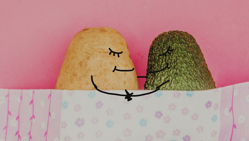 A yellow potato and a green avocado holding hands in bed with their eyes shut