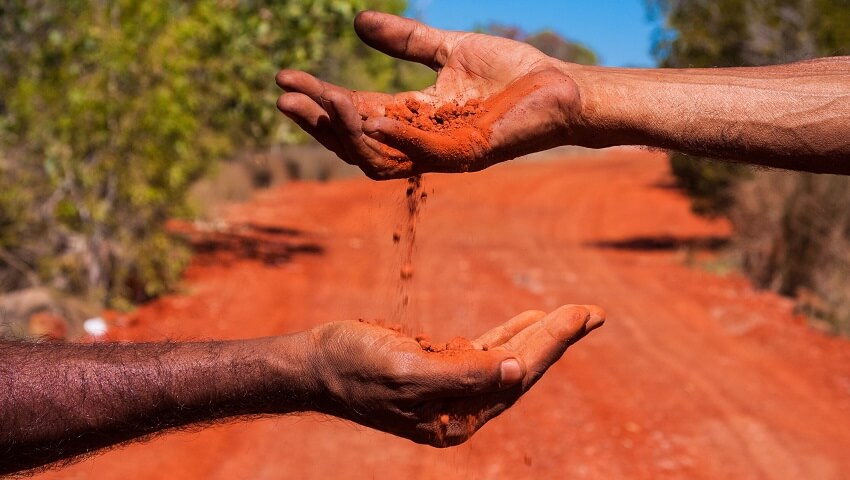Two hands against a red sand background. The top hand pours red sand into the bottom hand