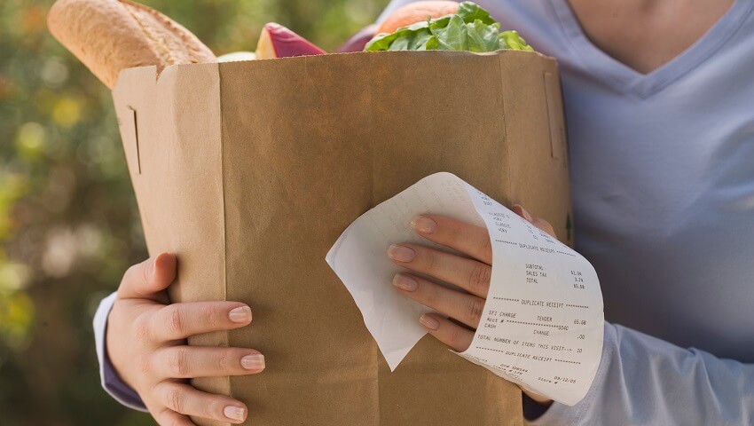 Woman holding a brown bag with bread and cabbage. She hold a paper that says 65 dollars