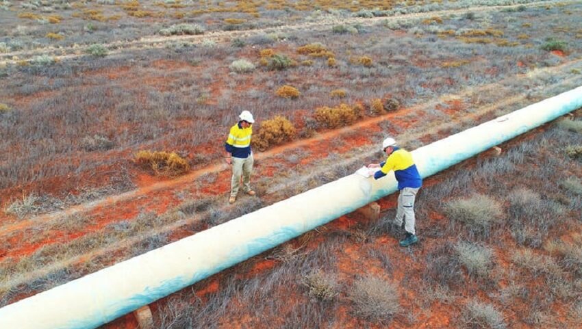 Two men stand next to a long white pipe. The ground is orange. They men are wearing yellow and blue sweaters. They wear white hats.