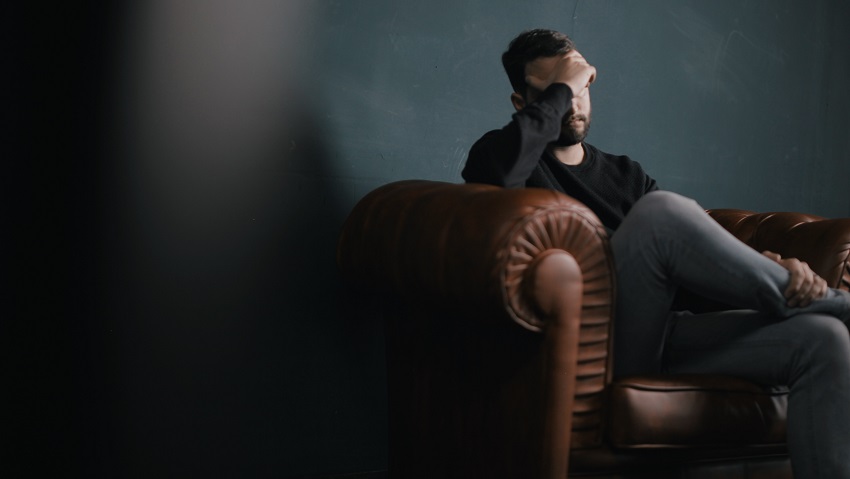 A man sitting on a brown couch. He has a hand over his face. The left side of the image is shadow