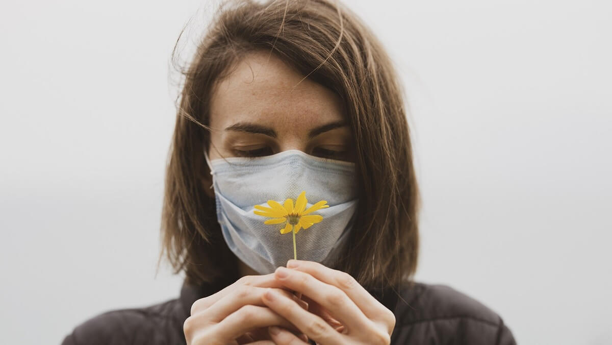 Woman with brown hair holds a flower to nose. SHe has a face mask on.