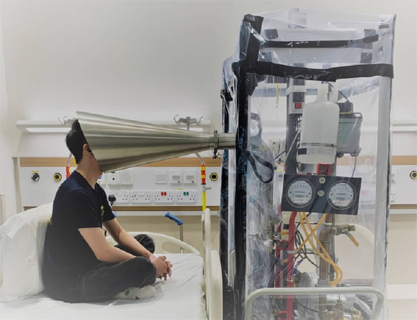 Man sits cross legged on a hospital bed with his face in a metal cone, connected to a large machine