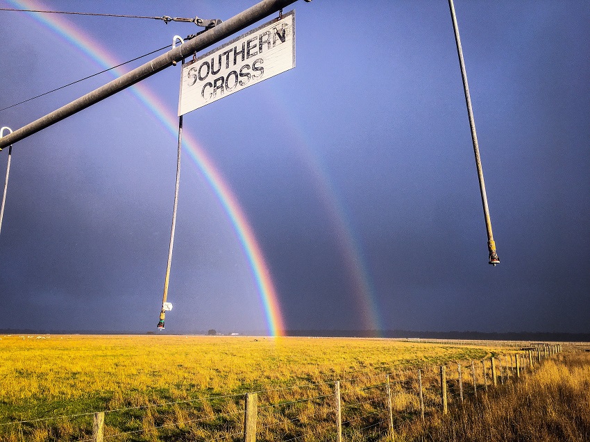 Road sign reading "southern cross" in front of a rainbow over a yellow field