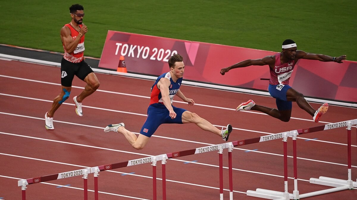 three men running. Thwo of them are jumping over hurdles. It says tokyo 2020 on the sideline