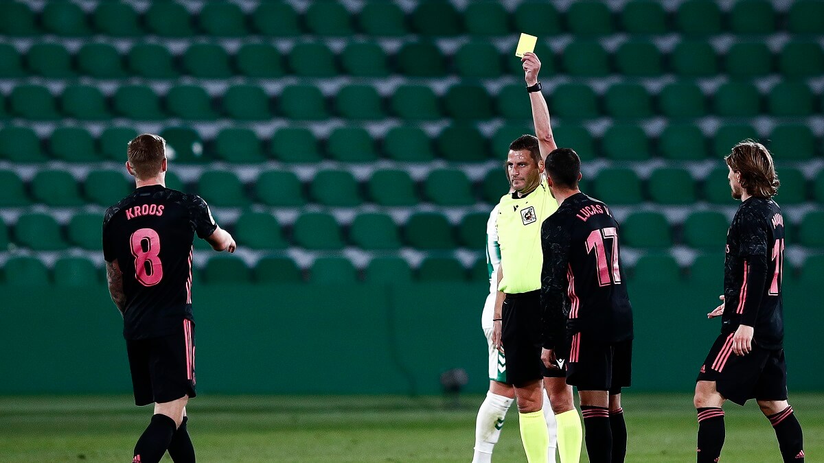 A man wearing yellow and black holds up a yellow card. Three men in black and pink look at him. Their are empty stadium seats in the background