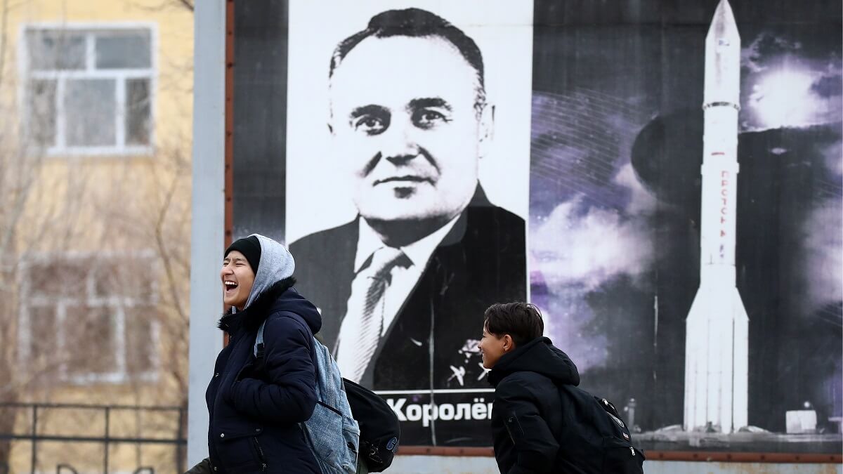 Children walk past a large poster of a man next to a rocket