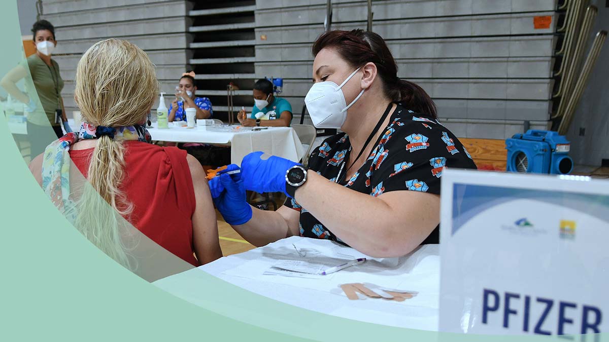 A woman wearing a mask with blue gloves. She is holding a needle and injecting another womans arm.