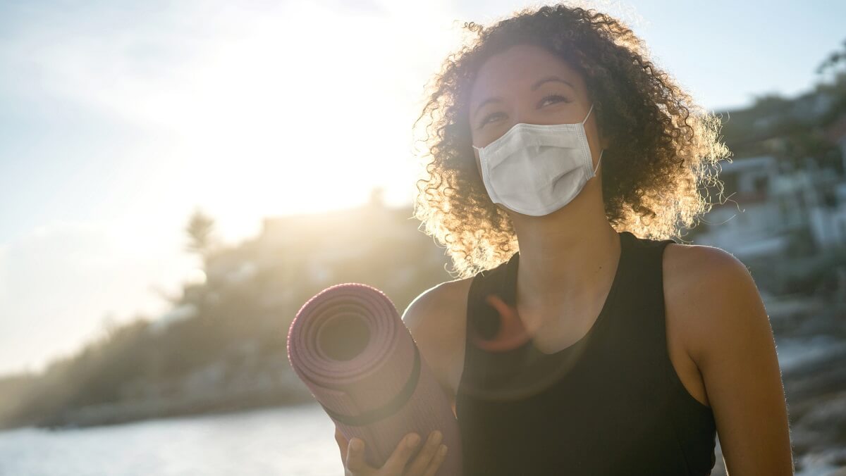 A woman holding an exercise mat with a facemask on