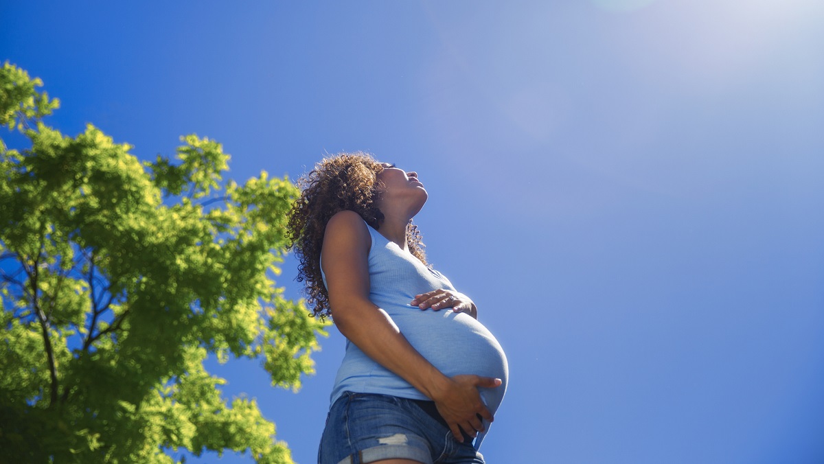 A pregnant woman holds her bump thoughtfully with a blue sky, trees, and sun flare behind her.