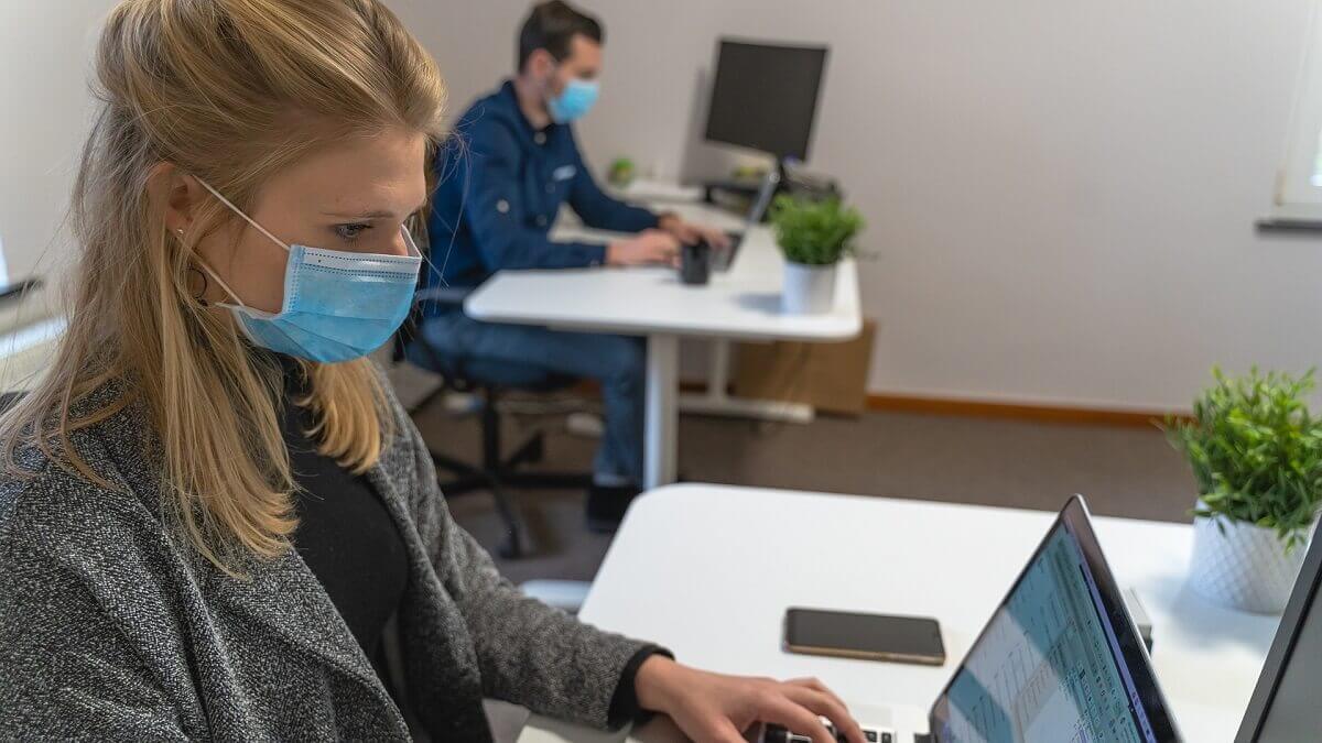 A woman sitting in an office with a laptop. She wears a mask