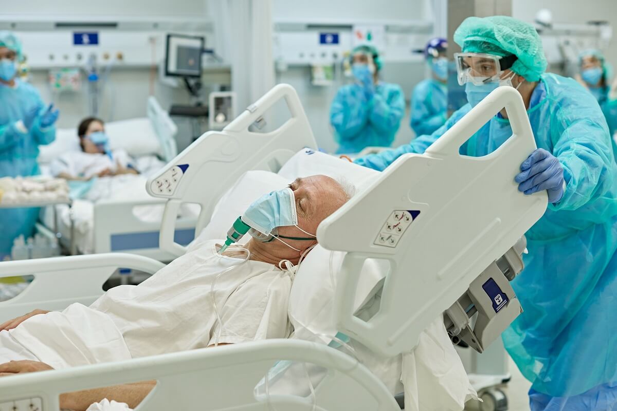 Senior caucasian man on ventilator lying in hospital bed with eyes closed as medical professional wheels his bed past camera.