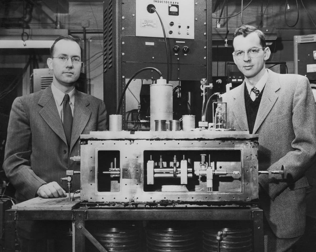 Black and white photo of two men in suits standing next to a device the size of a table