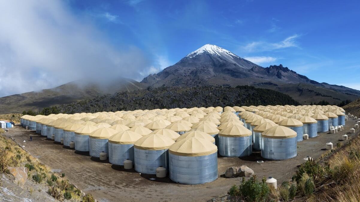 Lots of water tanks with cream coloured coverings outside. the sky is blue and there is a snow capped mountain in the background