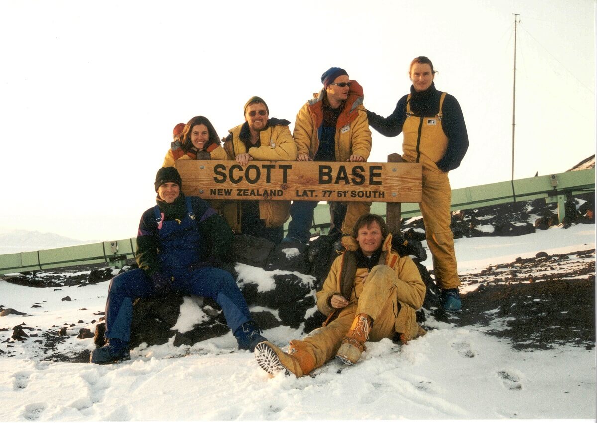 One woman and five men in polar outfits around a sign saying "scott base"