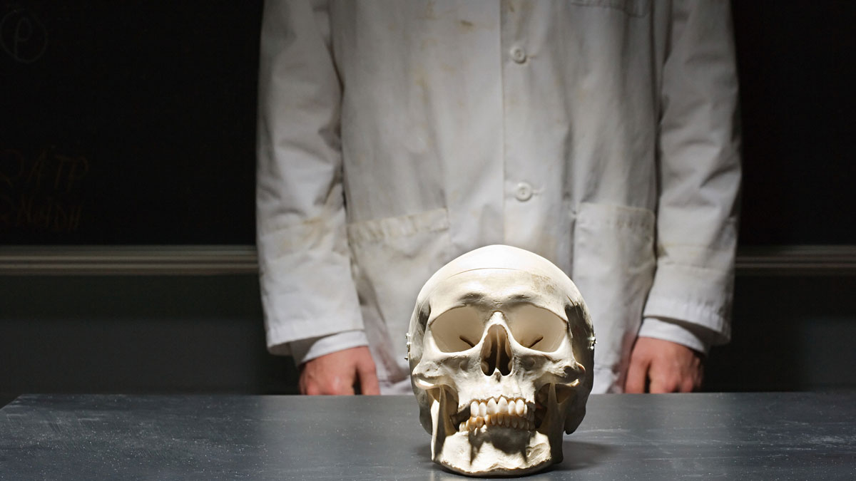 a person in a lab coat standing behind a human skull on a lab bench