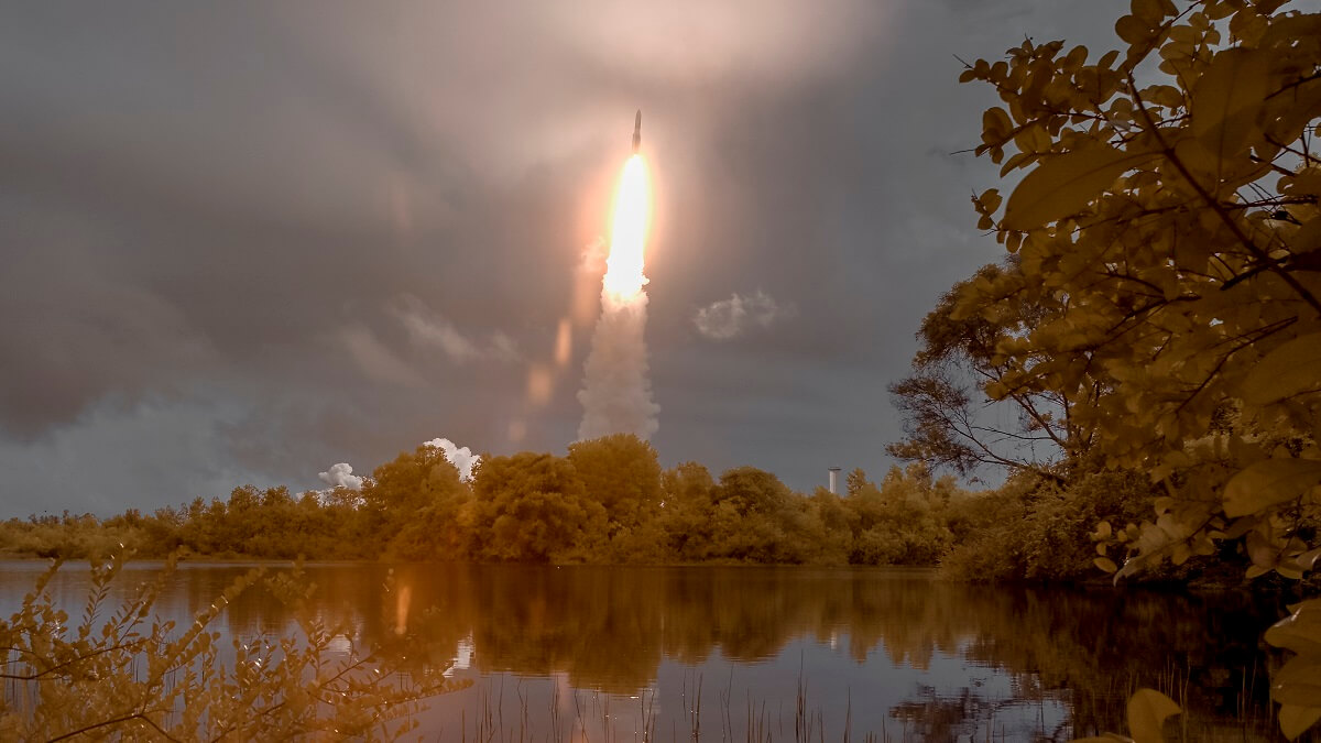 The James Webb Telescope launching into a grey sky, its blazing trail reflected in the waters of a lake.