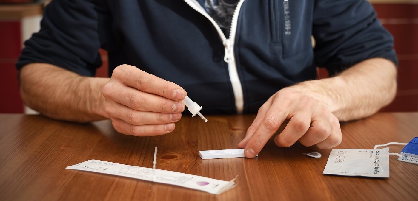 Man in a jacket sits at a table, using a dropper to apply a sample to a rapid antigen testing kit.