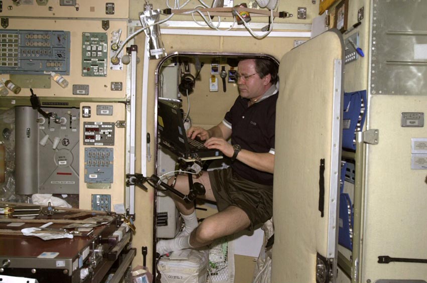 A man sitting in a small cupboard-like pod and using an old-fashioned computer on the international space station