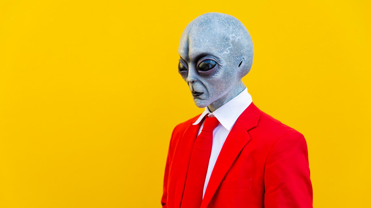 a stereotypical grey bald alien with large eyes wearing a red suit against an orange background