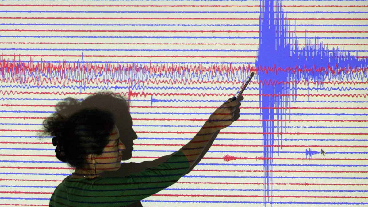 a woman pointing at blue and red seismograph lines projected onto a screen behind her