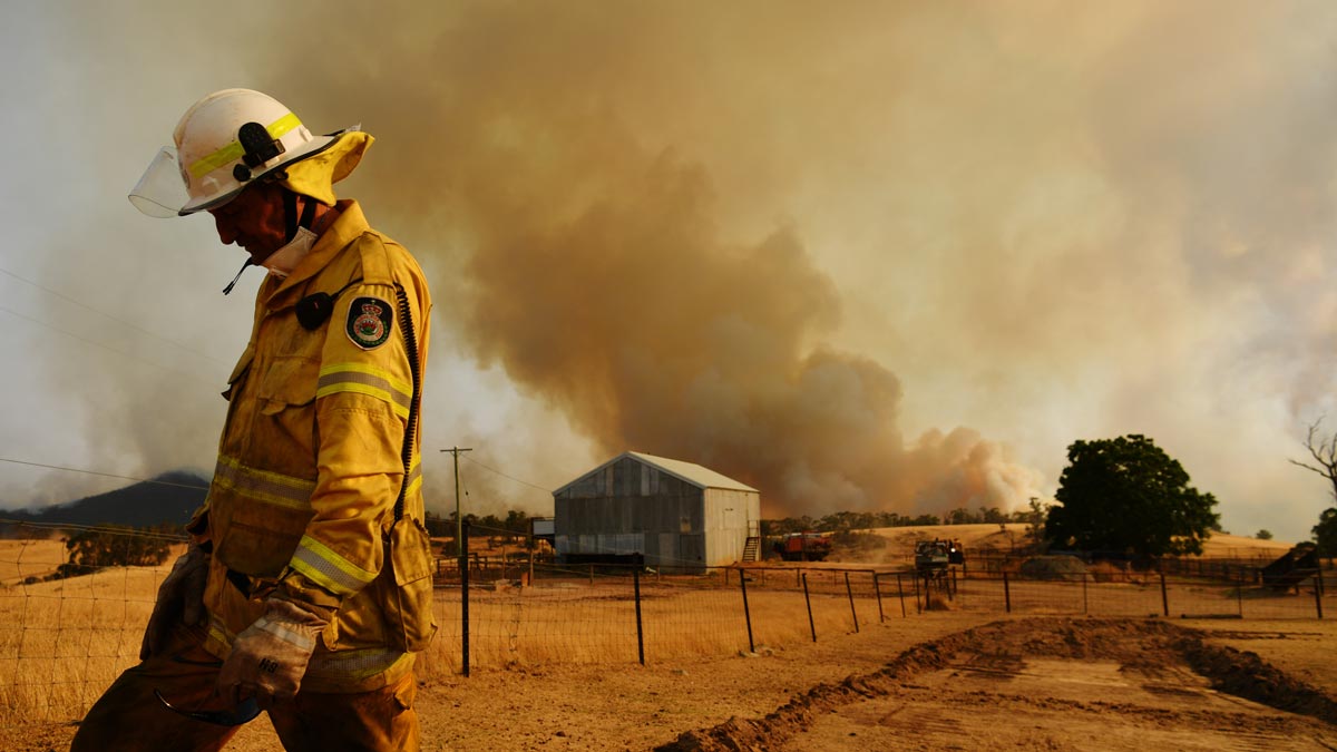 firefighter stands in the foreground in front of a tin shed and large plume of smoke from a bushfire