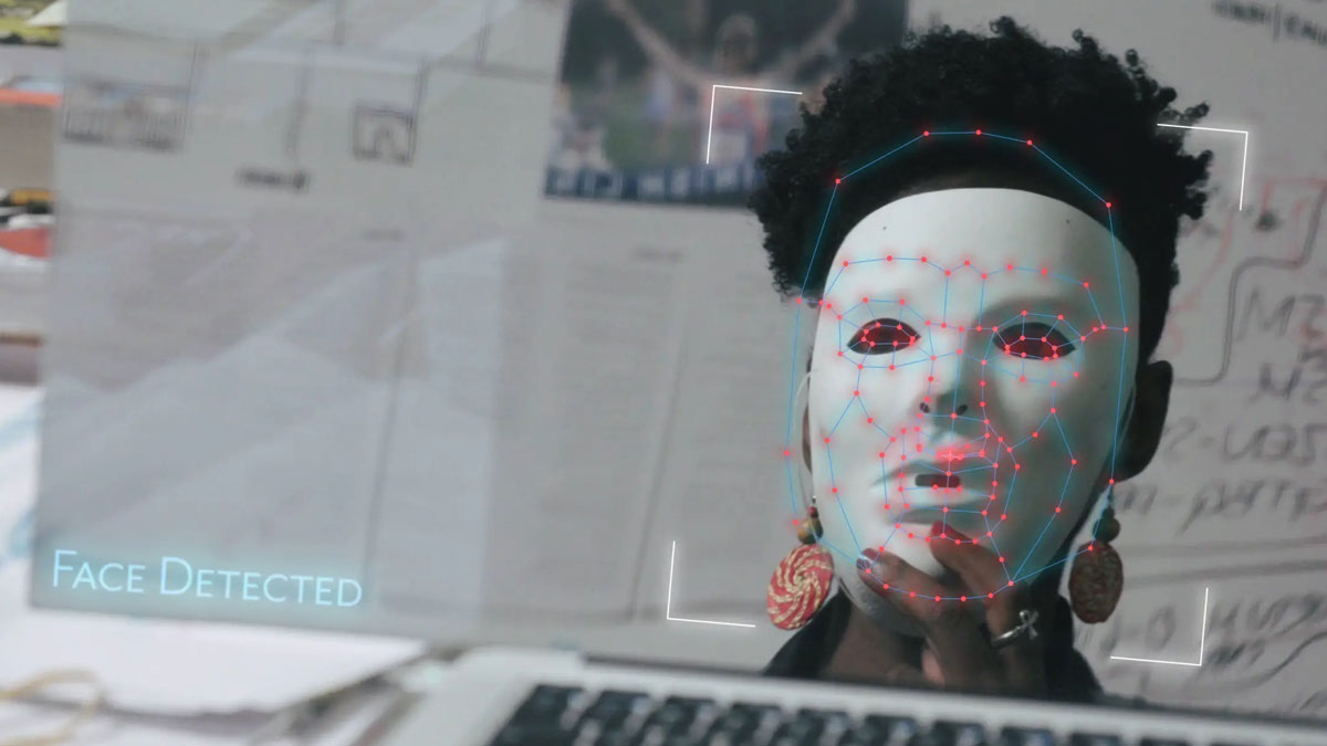 reflection of a dark-skinned woman in a computer screen who is holding up a white mask to her face the computer screen has a message saying face detected