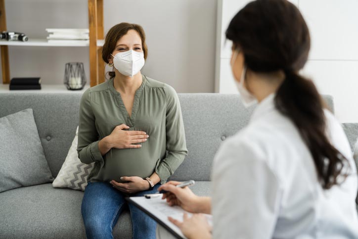 Pregnant woman wearing a face mask sits on a couch and speaks to a healthcare professional