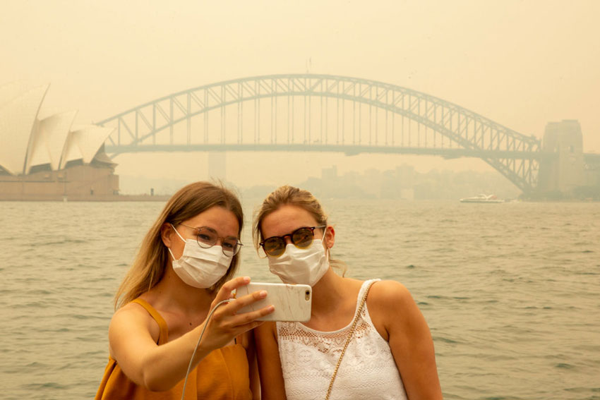 Two white women wearing surgical masks and taking a selfie in front of the sydney harbour bridge obscured by smoke from bushfires