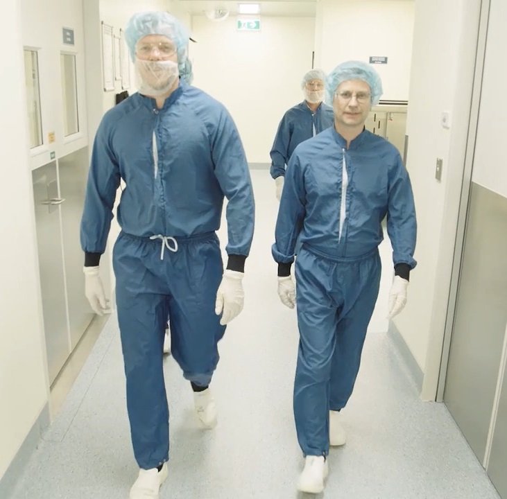 Four people walk down a corridor with blue coveralls, white shoes, white gloves, hair caps, beard nets and goggles