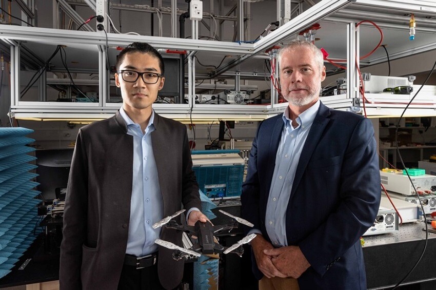 Two men stand in a lab, one man is holding a drone in his hand