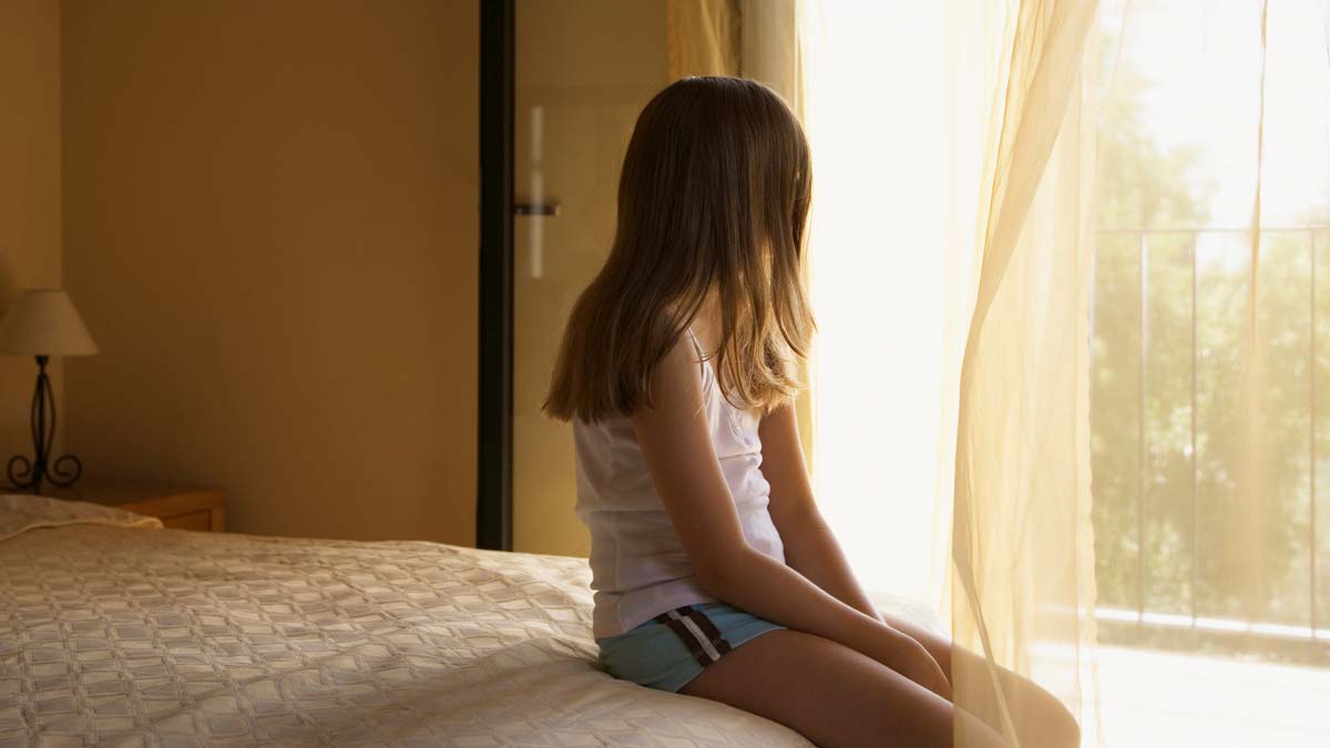 long covid in children concept a young girl in pyjamas sitting on a bed and looking out of a window