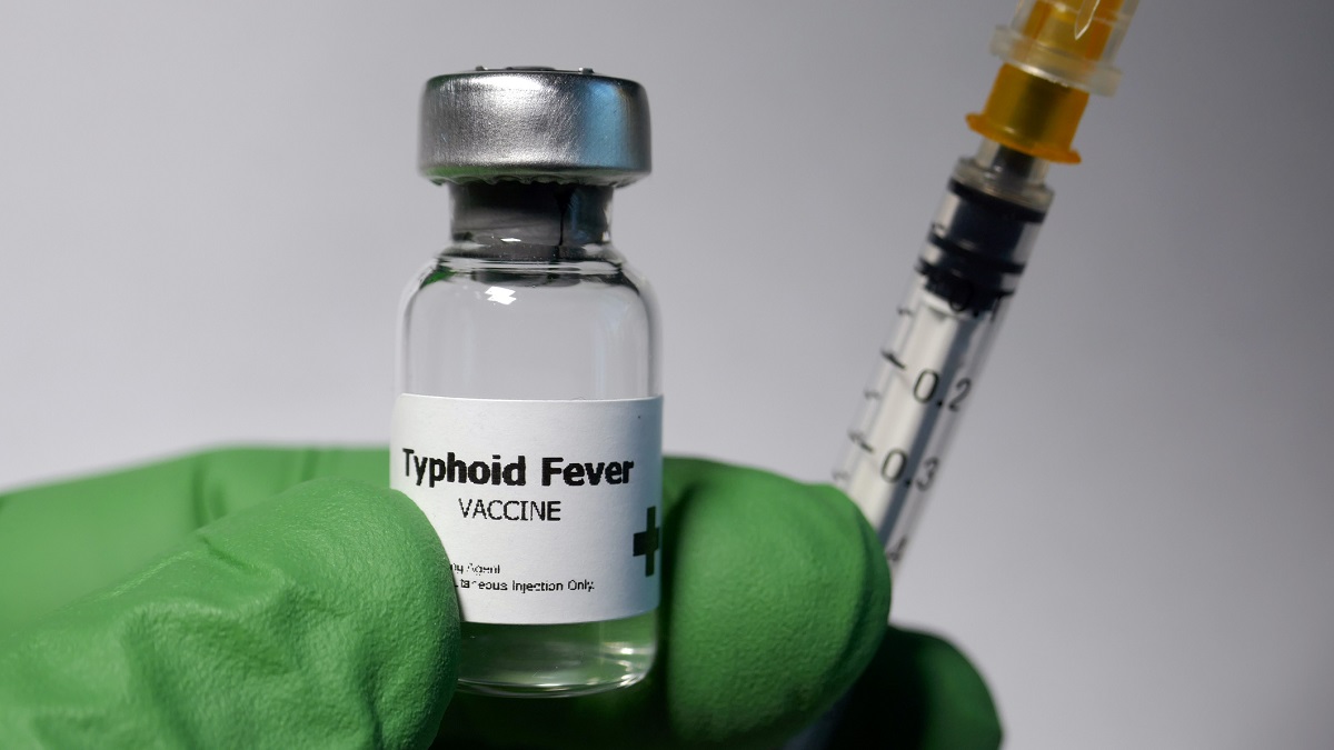 A hand wearing green gloves holds a vial labelled "Typhoid Fever Vaccine" and a needle
