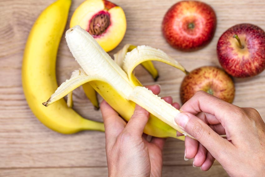 A person's hands peeling a banana with another banana, apples and nectarine in the background