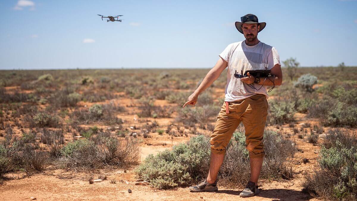 man stands in desert pointing to rock about the size of his finger while drone hovers in background