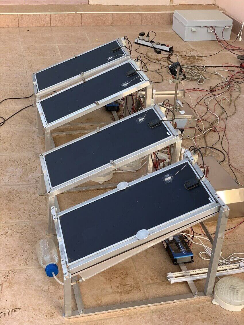Photo of four desk-sized solar panels connected to wires and metal frams, panel at the front has metal container below it and a tube running to a glass bottle