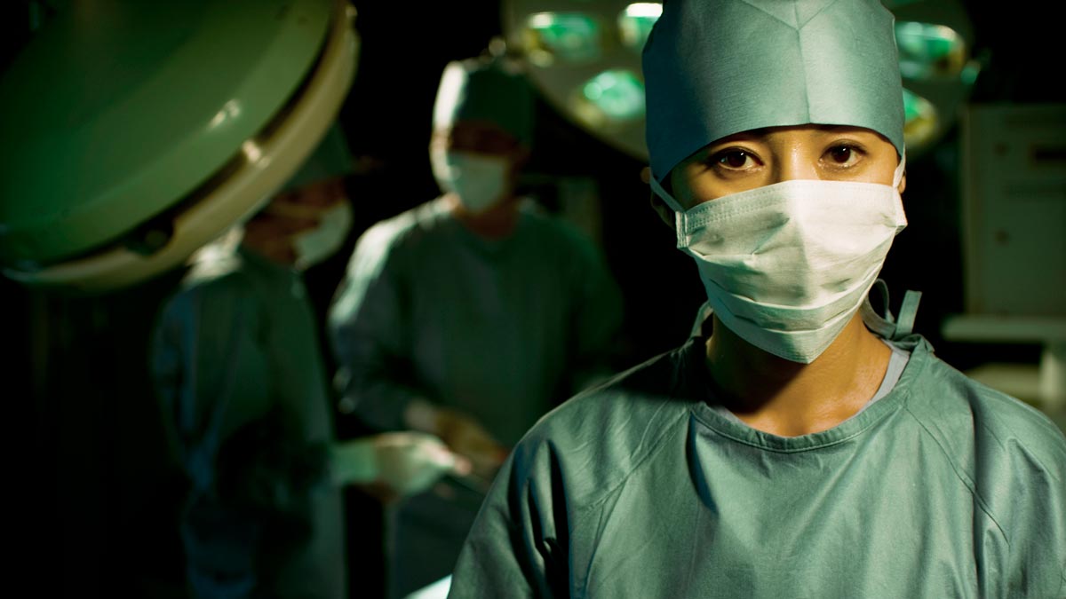 women in medicine concept a female surgeon wearing cap and mask looking at the camera with surgery in the background