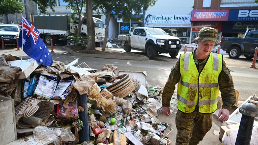 A white man wearing camouflage gear under a high-vis vest and walking past a pile of flood-damaged goods with an australian flag planted in it the main street and cars can be seen behind the man