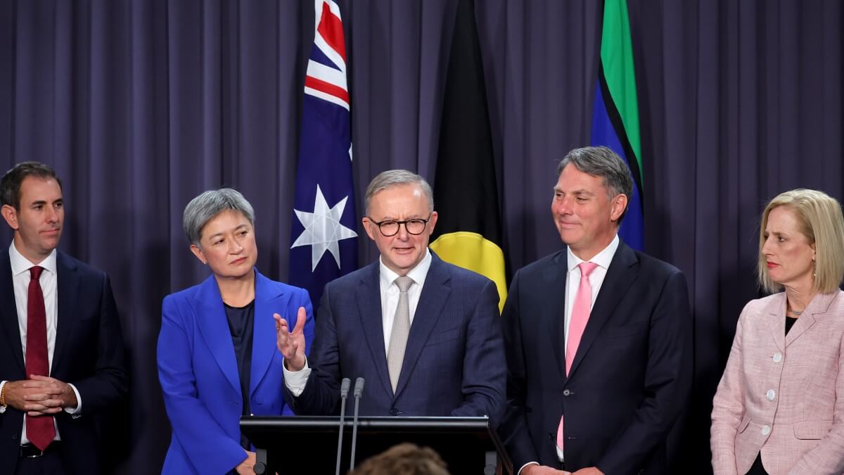 five people stand behind a lectern and in front of an Australian flag, an Aboriginal flag and a Torres Strait Island flag